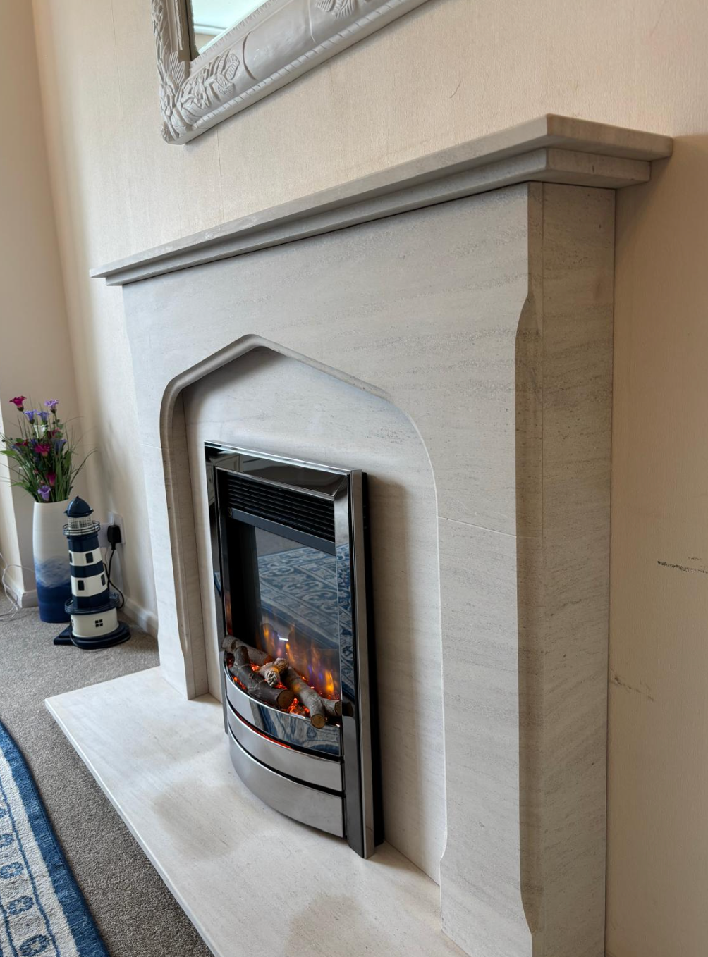 Cream fireplace with lit gas fire in a living room, beside a blue rug and a small decorative vase.