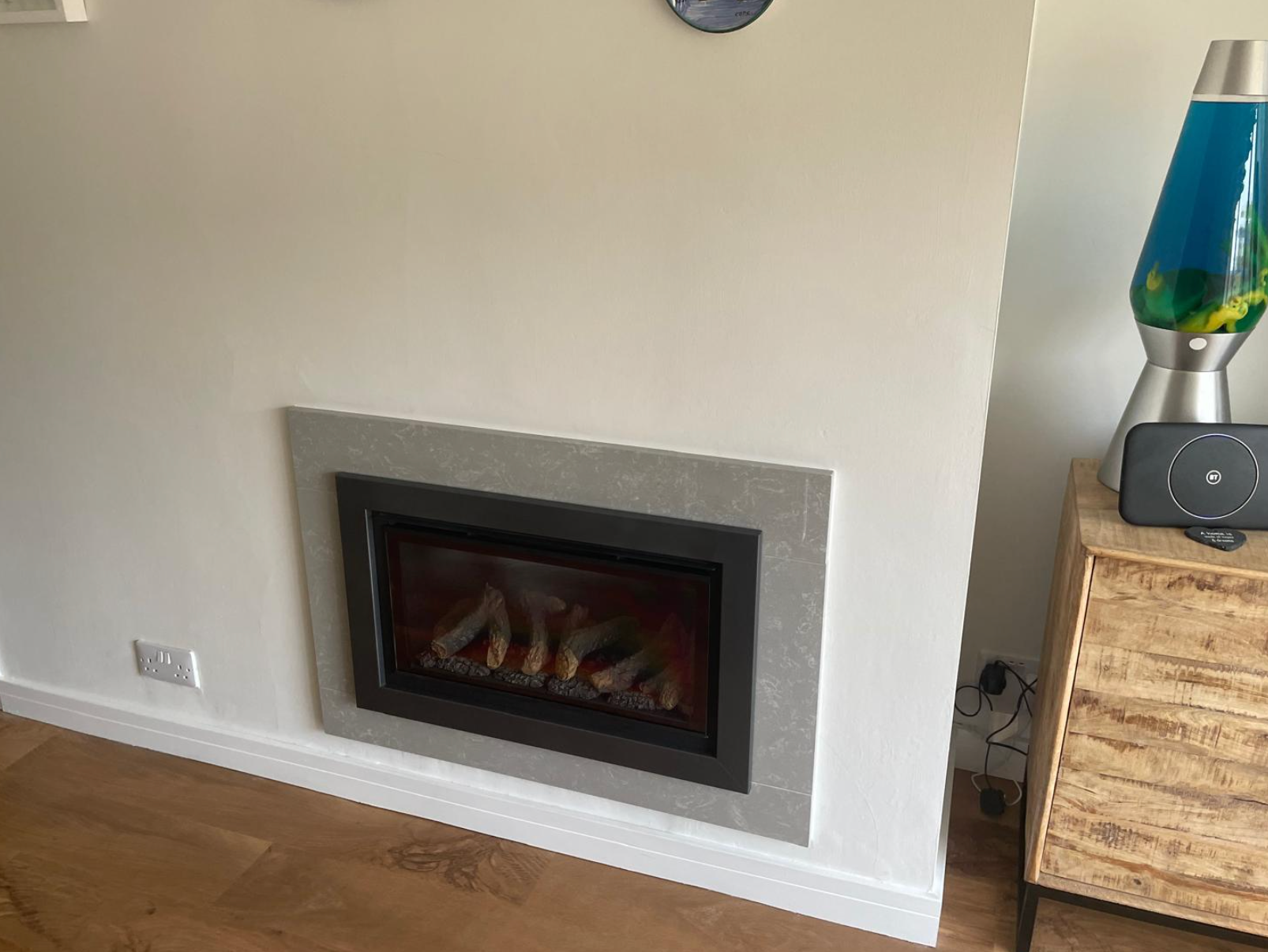 Modern wall-mounted fireplace with gray surround in a room with wood flooring and a lava lamp on a side table