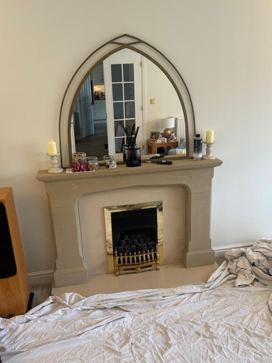 Bedroom with a beige fireplace and arched mirror, candles and decor on the mantel, and a bed in the foreground