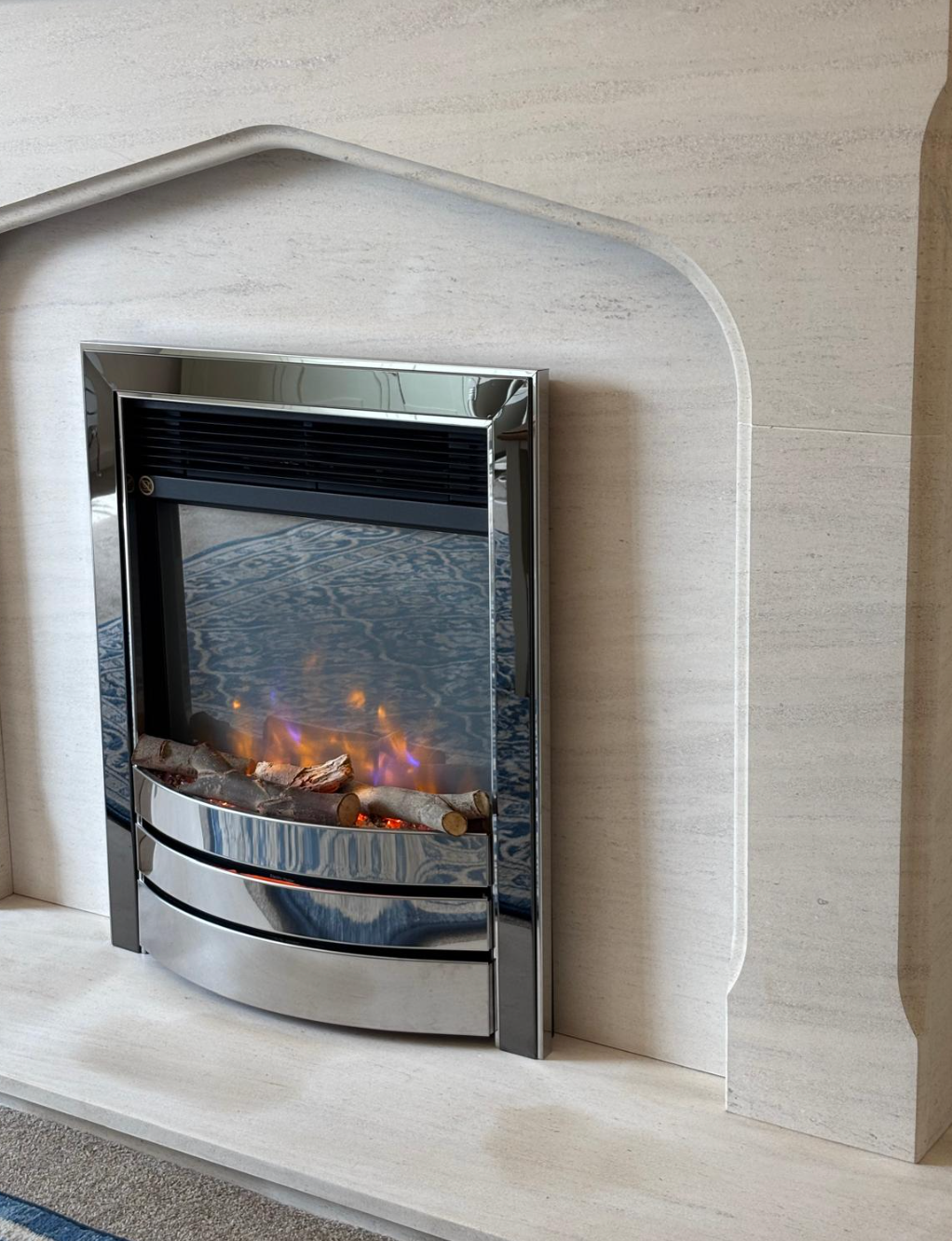 Modern silver electric fireplace inset in a beige wall, with glowing flames behind glass.
