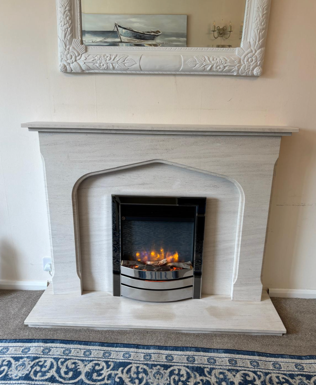 White fireplace with a lit electric fire beneath a framed picture on a beige wall.