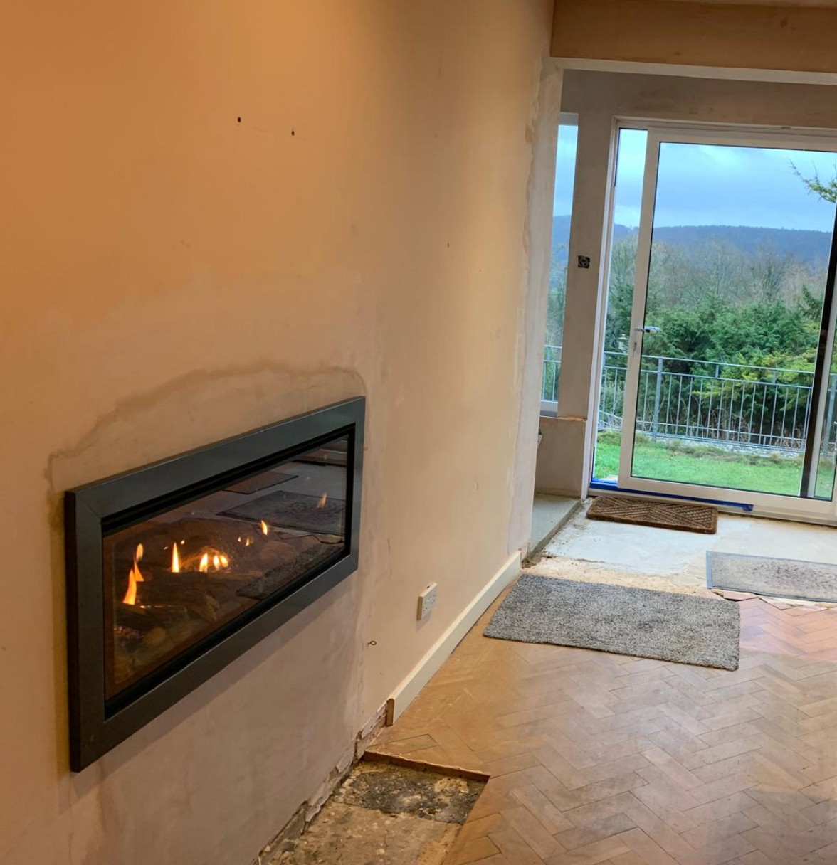 Modern living room with a lit wall-mounted fireplace and open glass doors to a deck and greenery outside