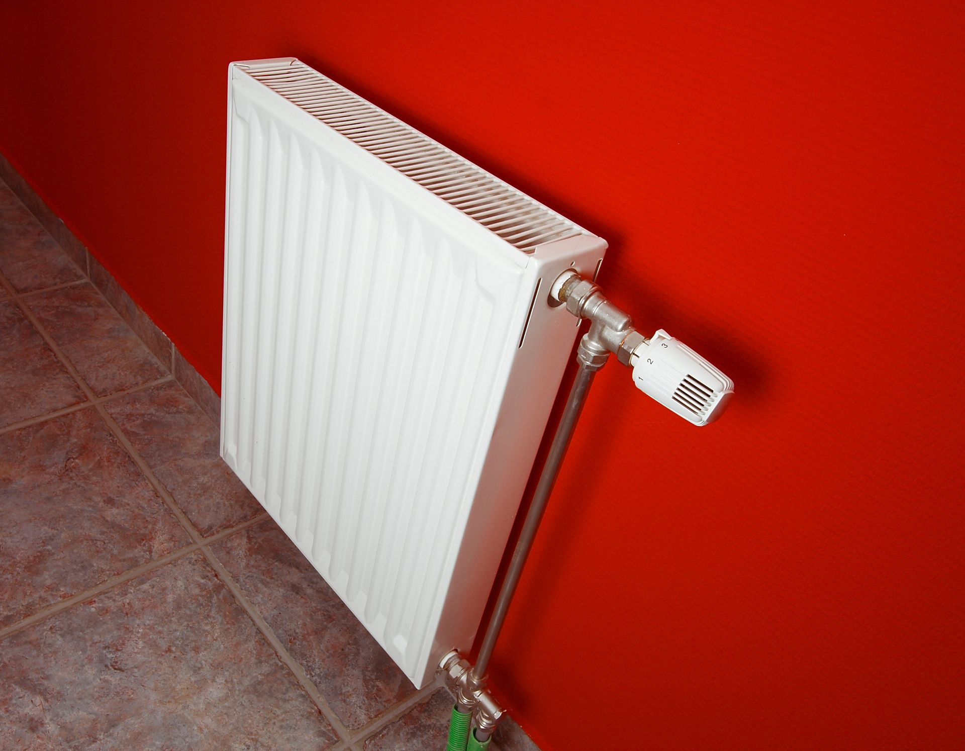 White radiator on a red wall above a tiled floor, with a valve on the right side.