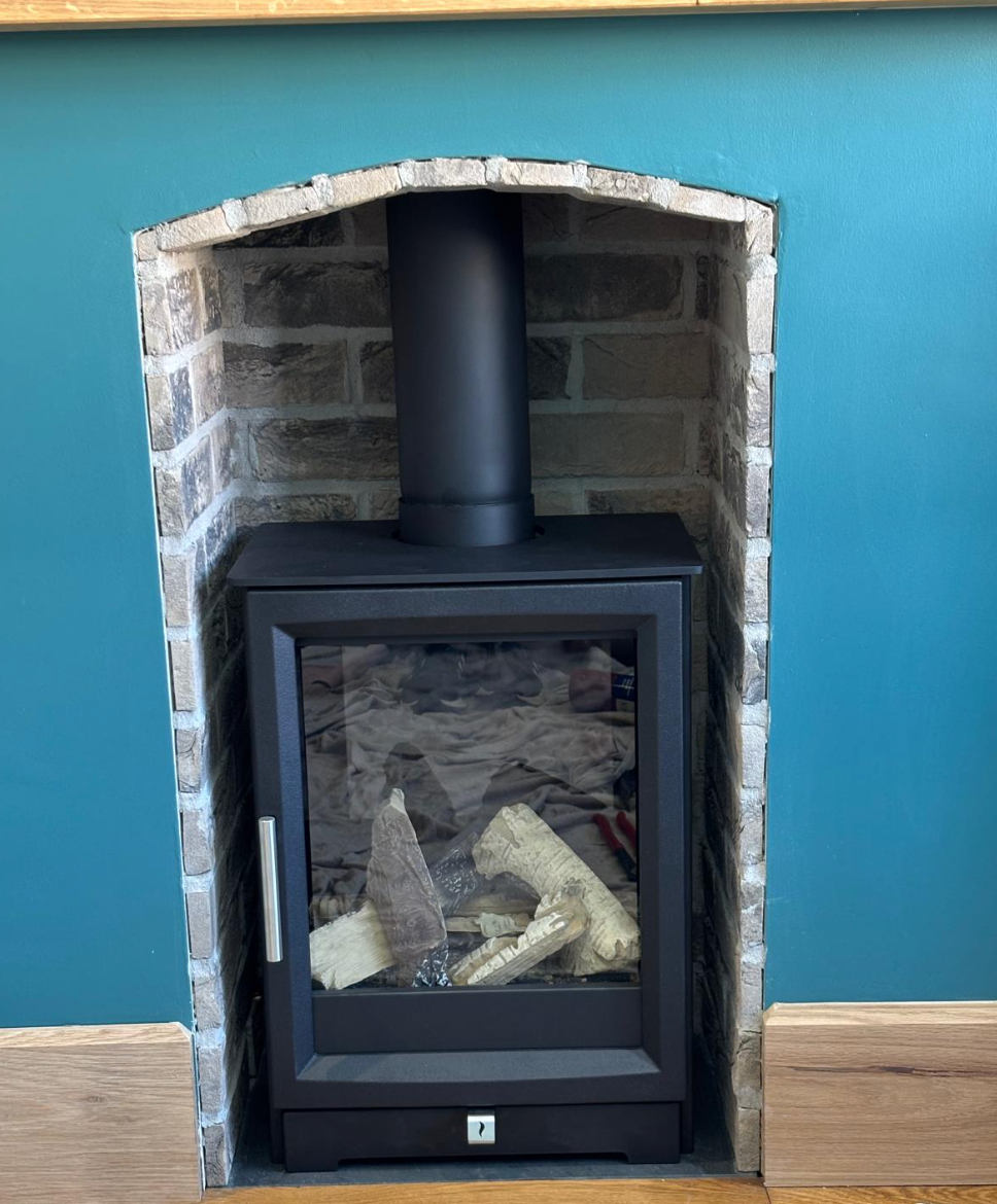 Black wood stove in a brick alcove against a teal wall, with logs inside and a chimney pipe above.