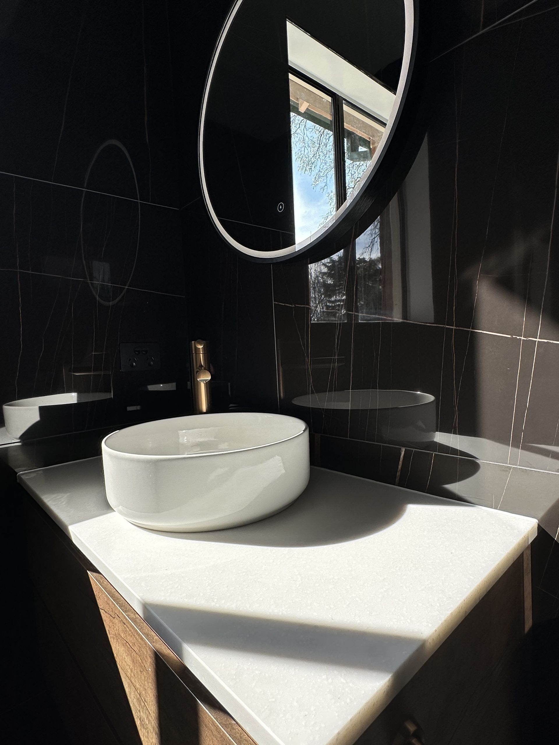 White sink on white countertop, black wall, round mirror, window — LD Carpentry in Goulburn, NSW