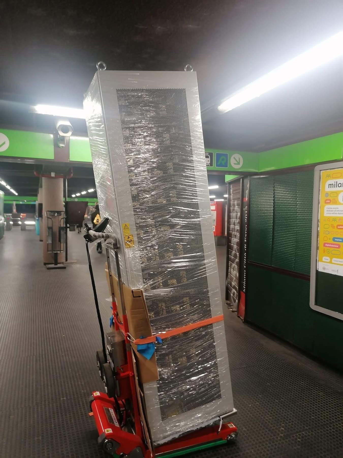Un elettrodomestico alto e imballato, posizionato su un carrello rosso, si trova in un corridoio interno con pareti verdi e luci intense sul soffitto.