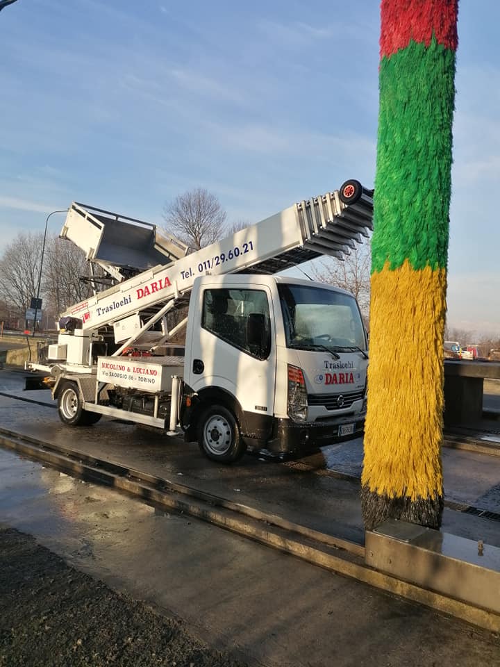 Un camion con piattaforma elevatrice bianca lava un alto palo a strisce a lato di una strada bagnata, sotto un cielo sereno.
