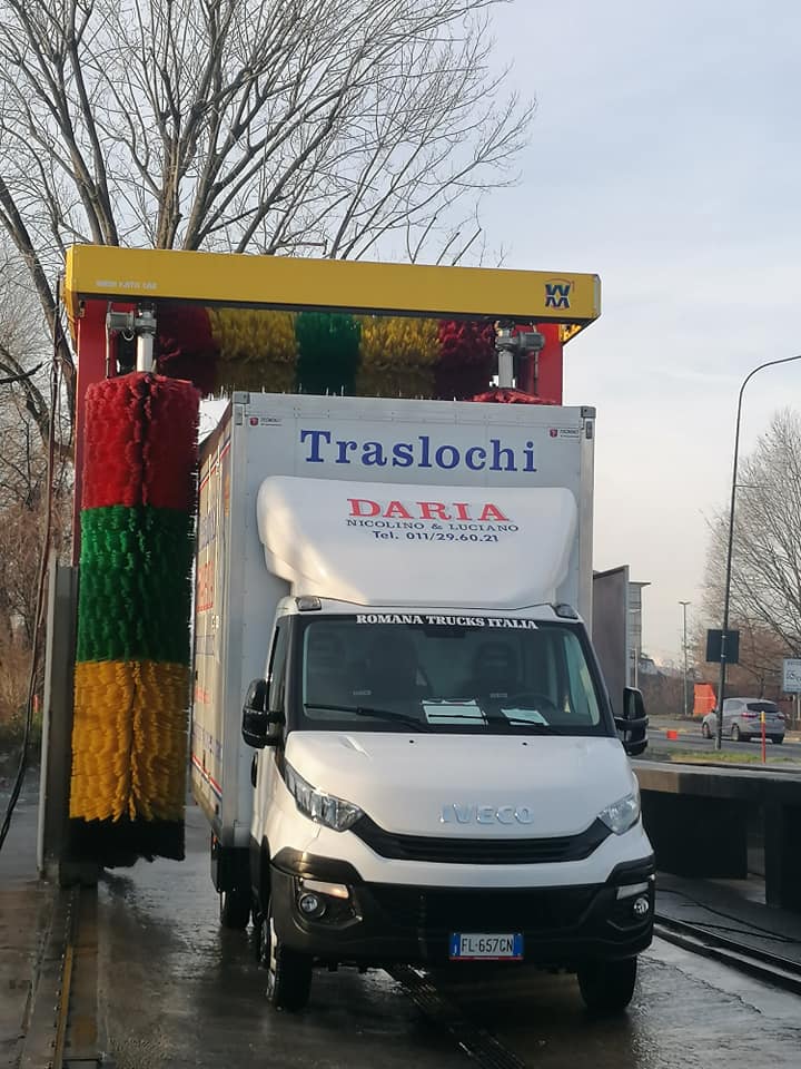 Un camioncino bianco per traslochi sotto un colorato arco di lavaggio su una strada bagnata, con la scritta
