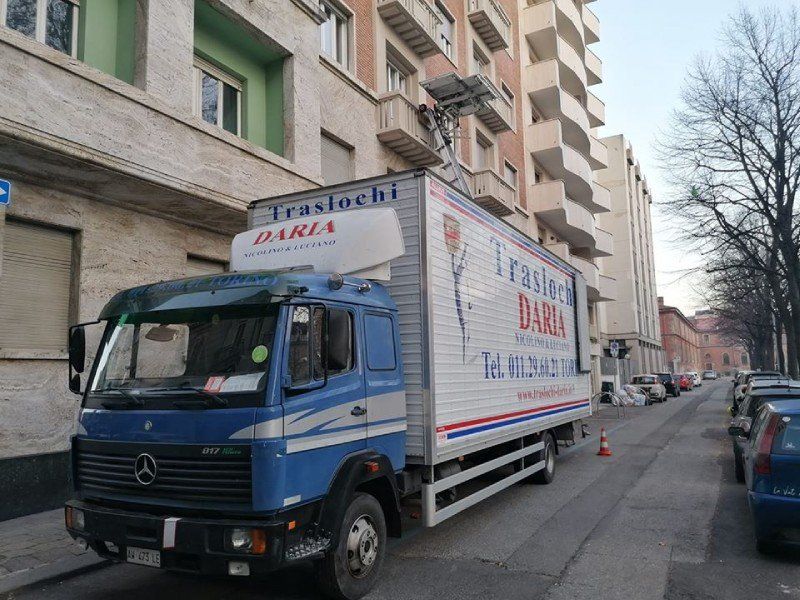 Un camion per traslochi Mercedes blu parcheggiato in una stretta via cittadina accanto a dei palazzi residenziali.