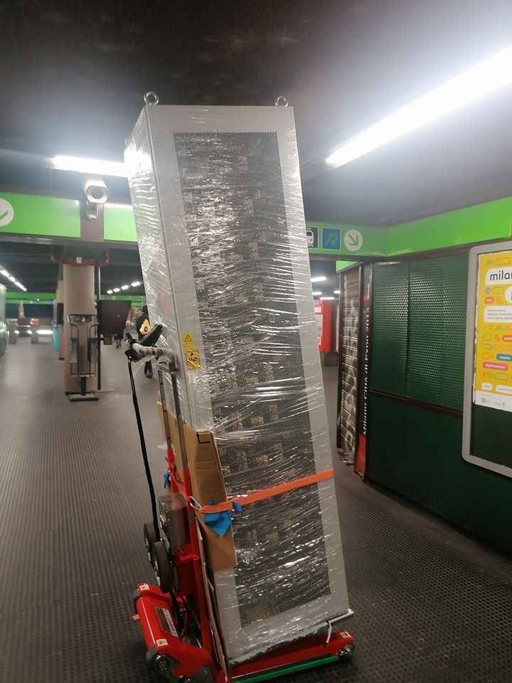 Un elettrodomestico alto, avvolto in pellicola termoretraibile, su un carrello rosso, si trova nel corridoio di una stazione con pareti verdi e luci brillanti.