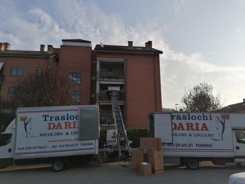 Un camion per traslochi con una scala all'ingresso di un edificio, scatole sul marciapiede e un secondo camion nelle vicinanze.