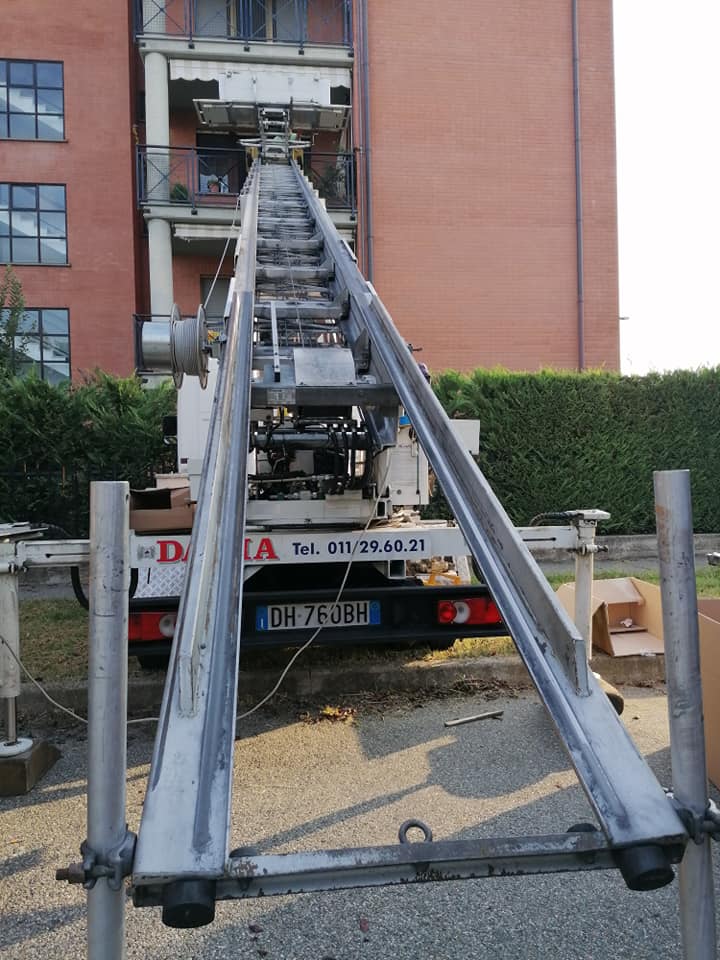 Due lunghe travi metalliche che si estendono da un camion bianco in un cantiere all'aperto.