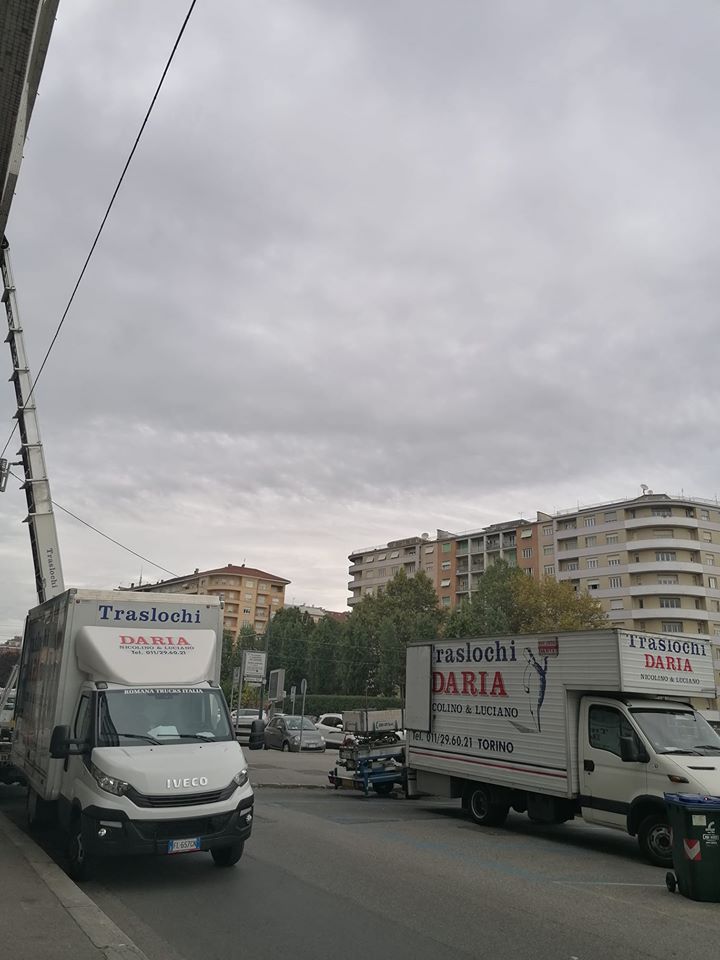 Due camion per traslochi su una strada cittadina nuvolosa, accanto a palazzi e linee elettriche.