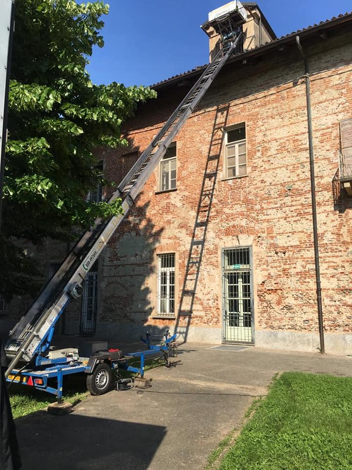 Due scale appoggiate a un edificio in mattoni, con un ascensore blu parcheggiato sotto e un albero accanto.
