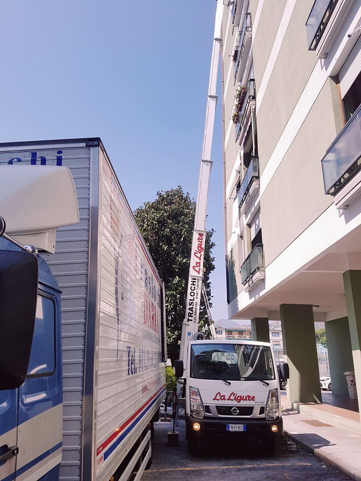 Due camion su una strada stretta accanto a un edificio, con un piccolo camion bianco rivolto in avanti davanti a un camion più grande.