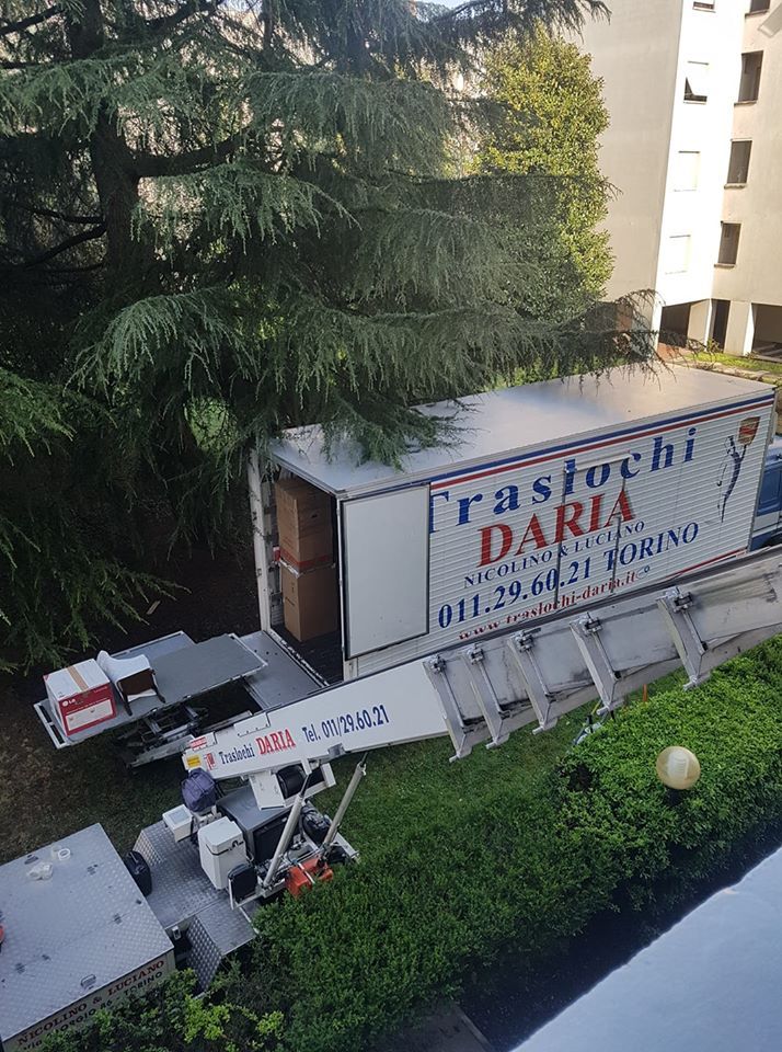 Un camion per il trasporto di pianoforti è parcheggiato vicino a un edificio, con gli addetti che caricano l'attrezzatura accanto a una siepe.