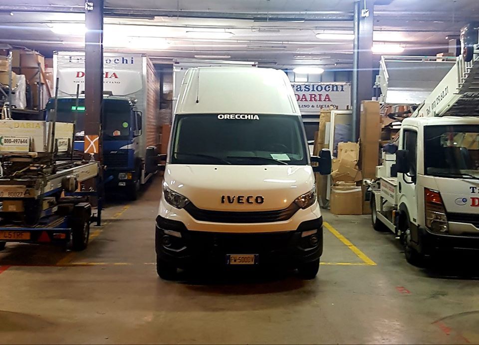 Un furgone Iveco bianco è posizionato al centro di un garage ben illuminato, affiancato da altri camion e attrezzature.