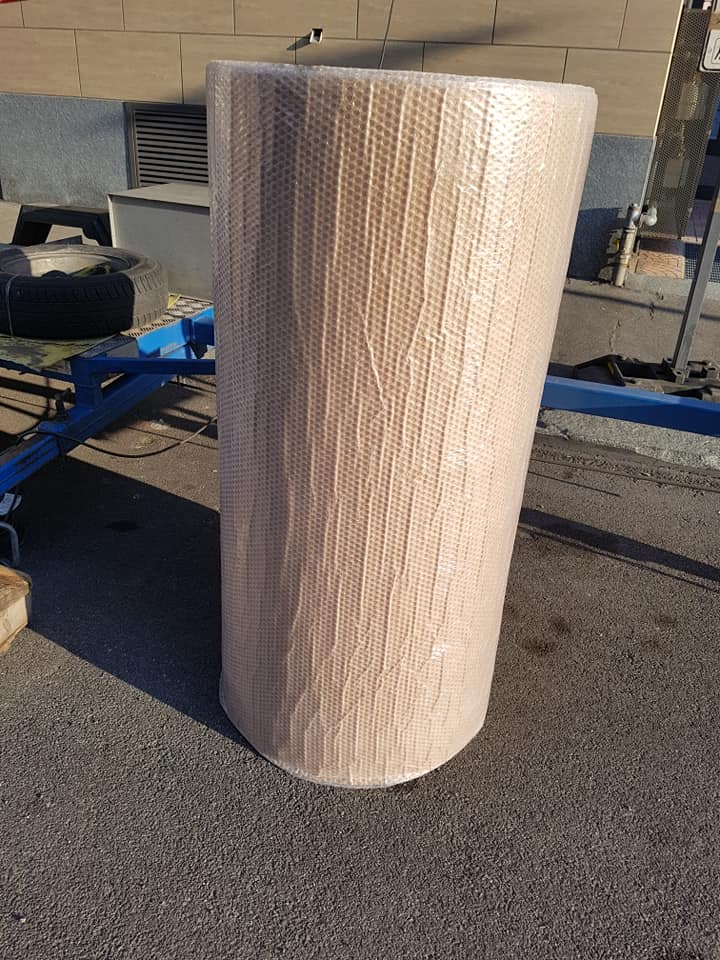 Un grande rotolo cilindrico bianco avvolto nella pellicola trasparente, appoggiato all'aperto sul marciapiede vicino a un carrello blu e al muro di un edificio.