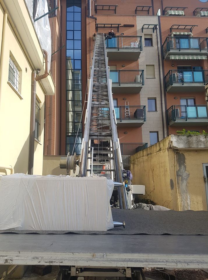 Una lunga scala appoggiata a un condominio, con operai sul tetto e un oggetto ricoperto di bianco in primo piano.