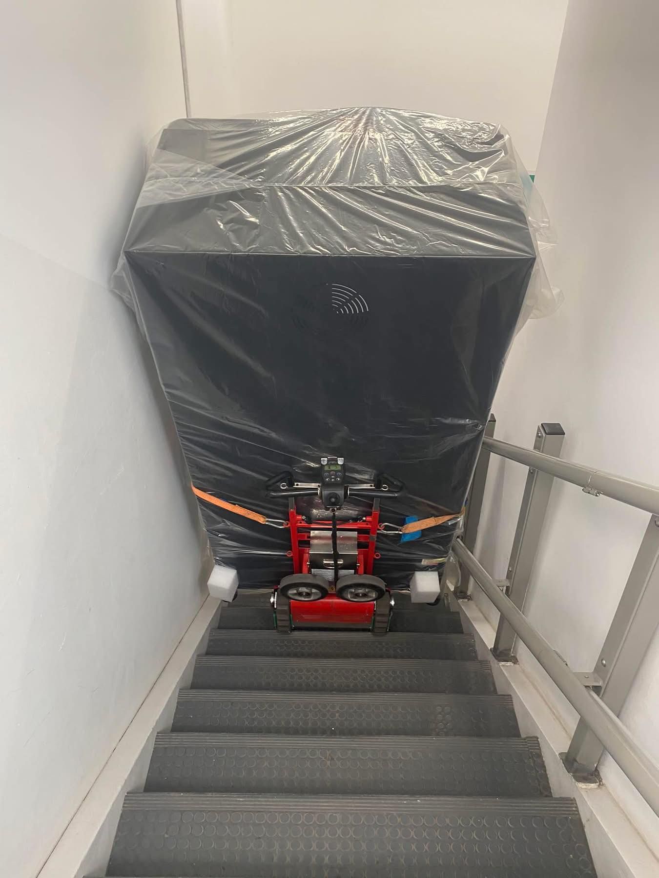 Un grosso oggetto avvolto in un telo nero ostruisce una stretta tromba delle scale, con un carrello a mano rosso in fondo.