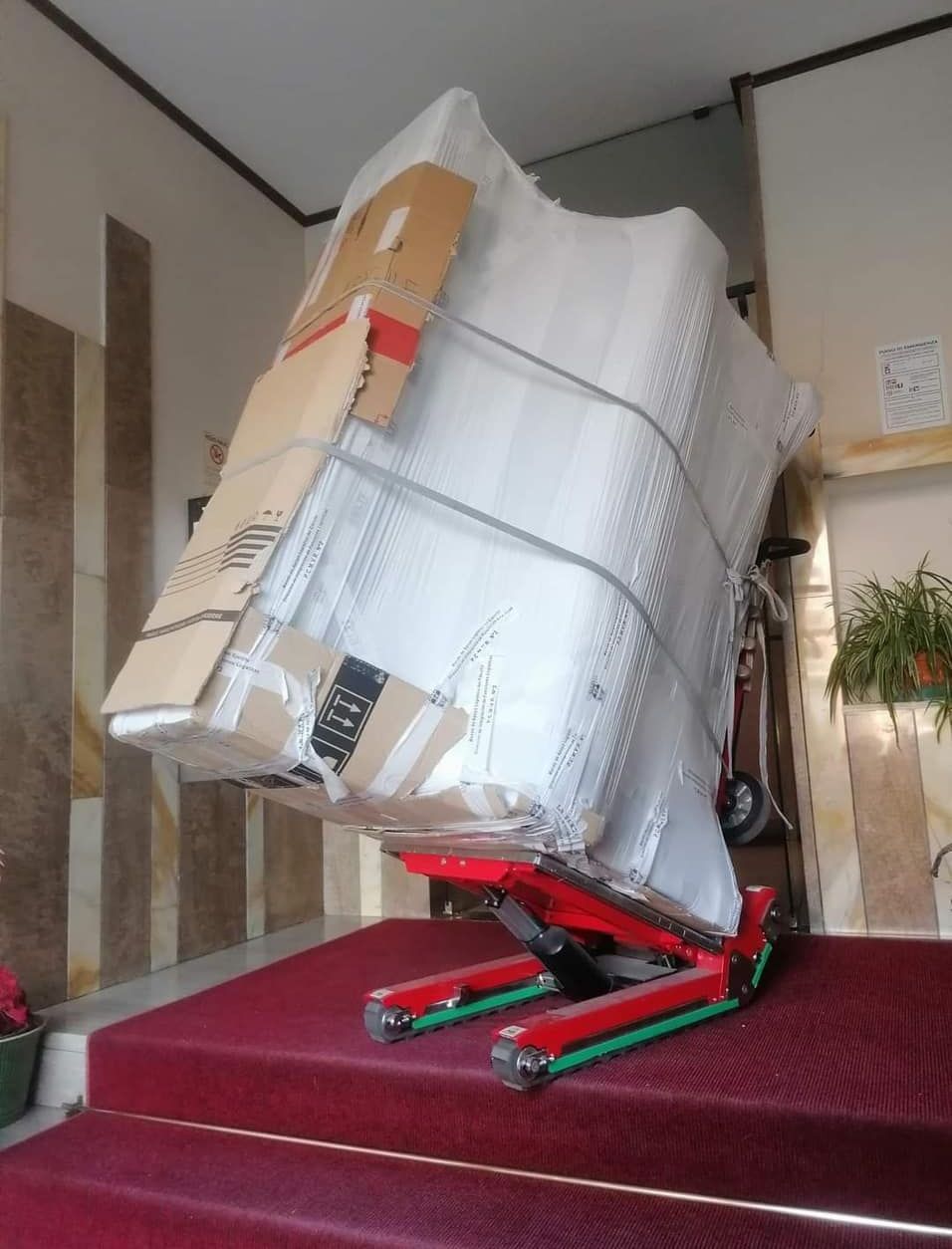 Un grosso oggetto bianco, imballato in un pacco, è fissato a un telaio rosso con ruote su un pianerottolo interno ricoperto di moquette.