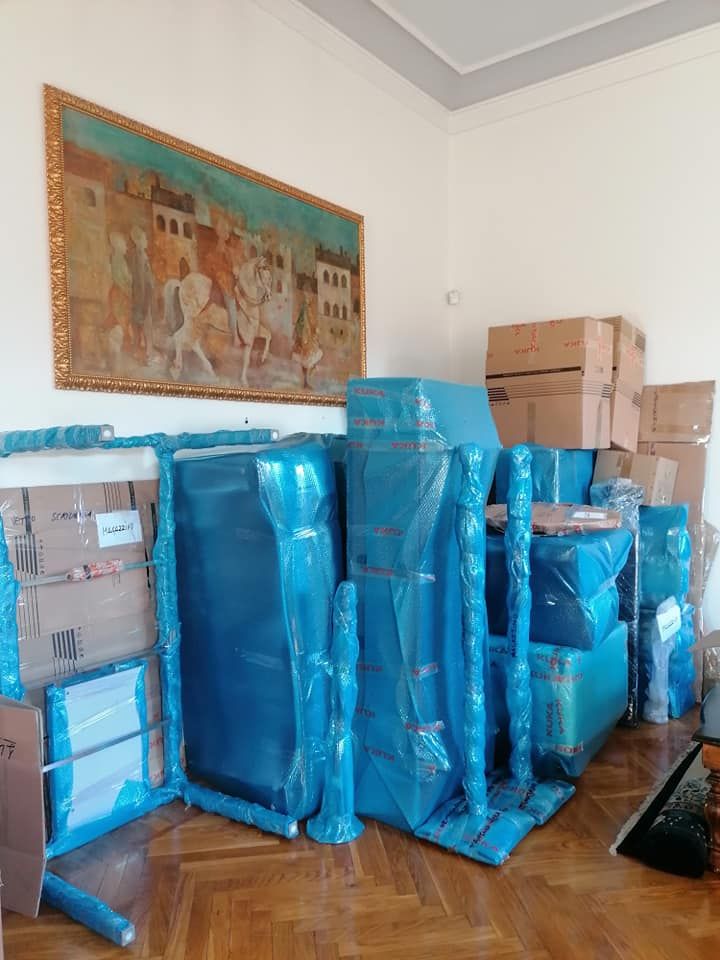 In una stanza con un quadro appeso, si trovano scatole di cartone impilate e mobili avvolti in plastica blu.