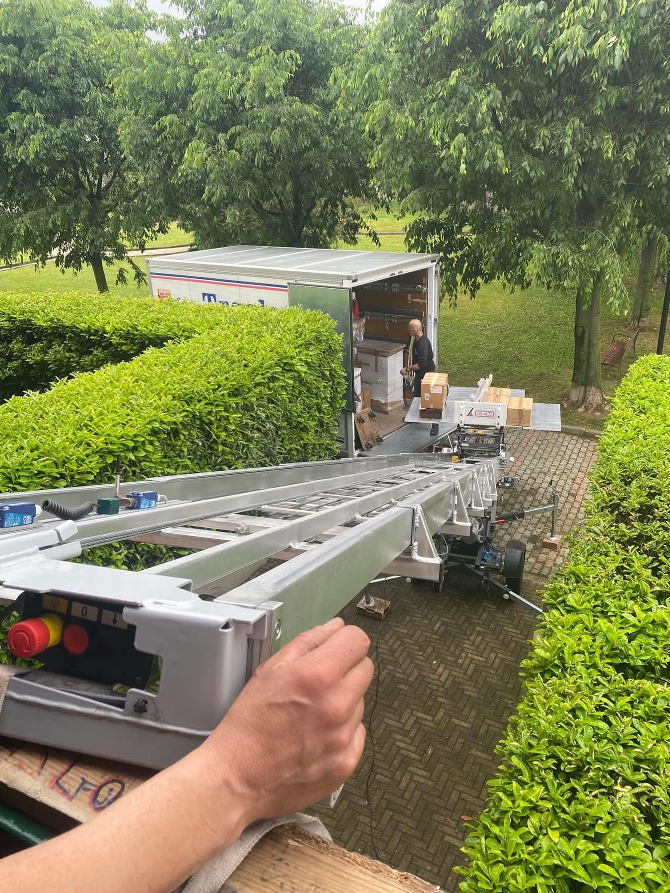 Scala montata su un camion che si estende attraverso una siepe in un giardino, con operai e scatole nelle vicinanze.