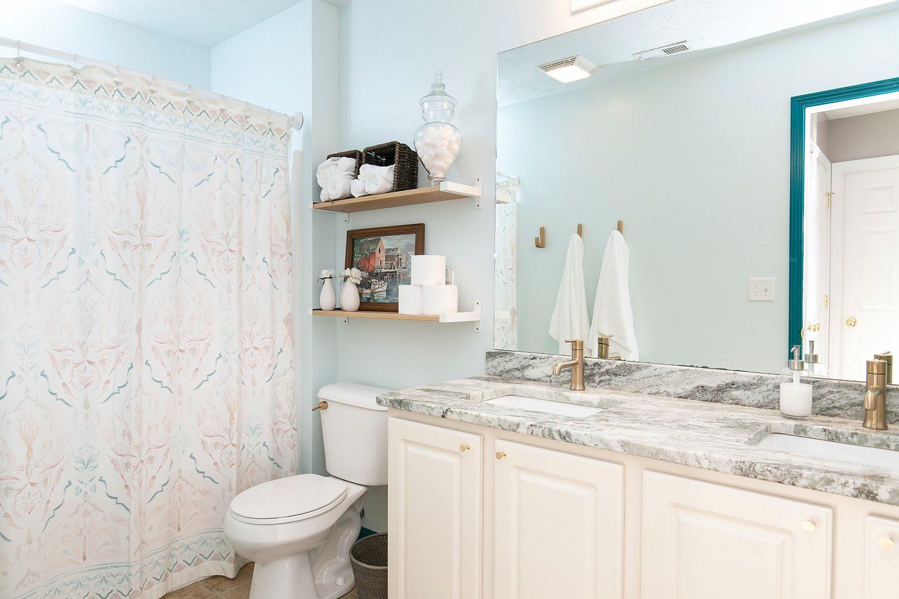 Bathroom with toilet, white vanity, marble countertop, and shower curtain. Light blue walls and decorative shelves.