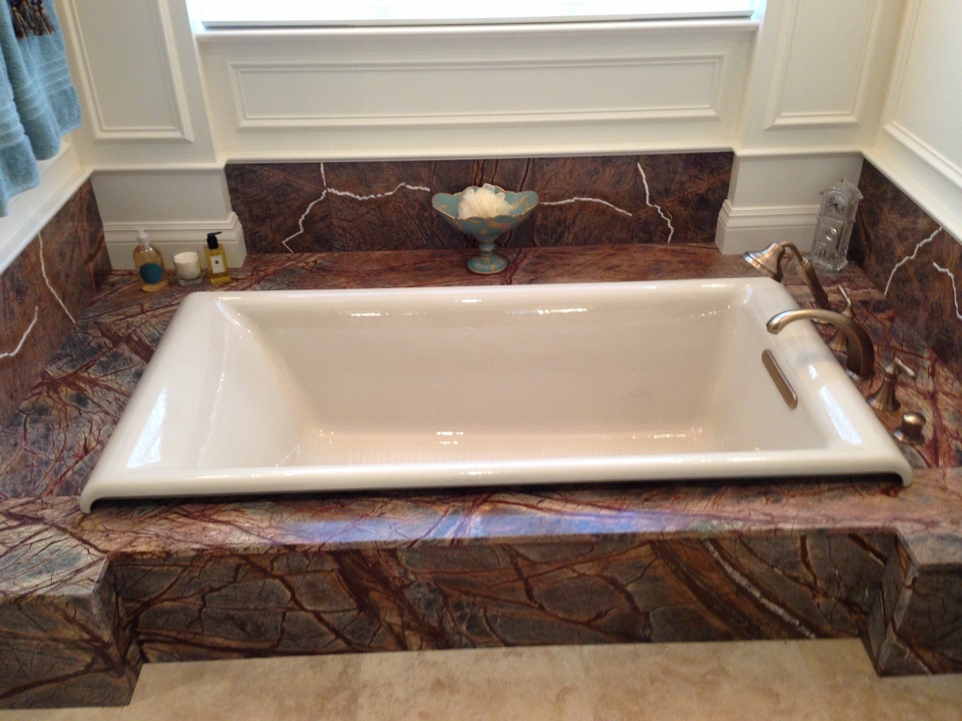 A built-in white bathtub surrounded by brown and cream marble, under a window with a shelf.