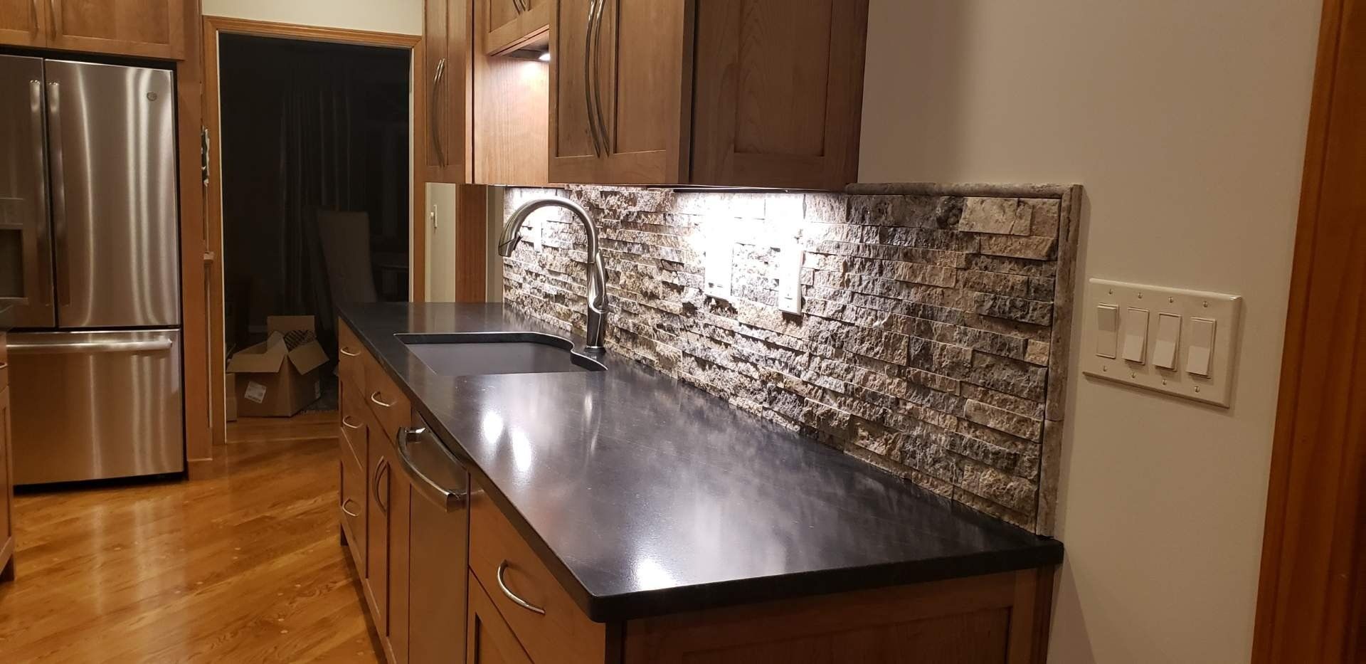 Modern kitchen with dark granite counters, wood cabinets, stainless fridge, and tile backsplash; warm lighting.