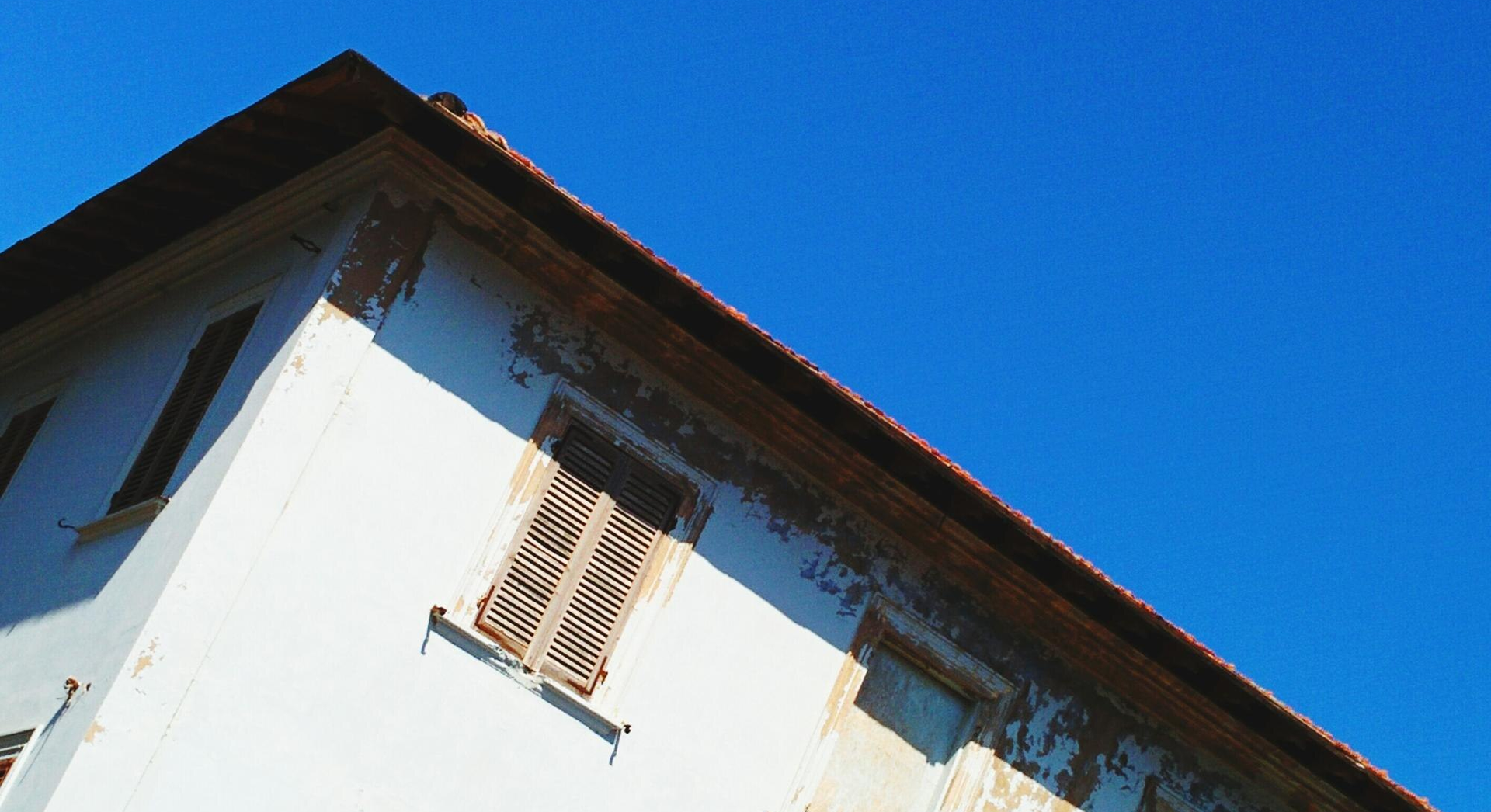 Corner of a white building with peeling paint, wooden shutters, against a bright blue sky.