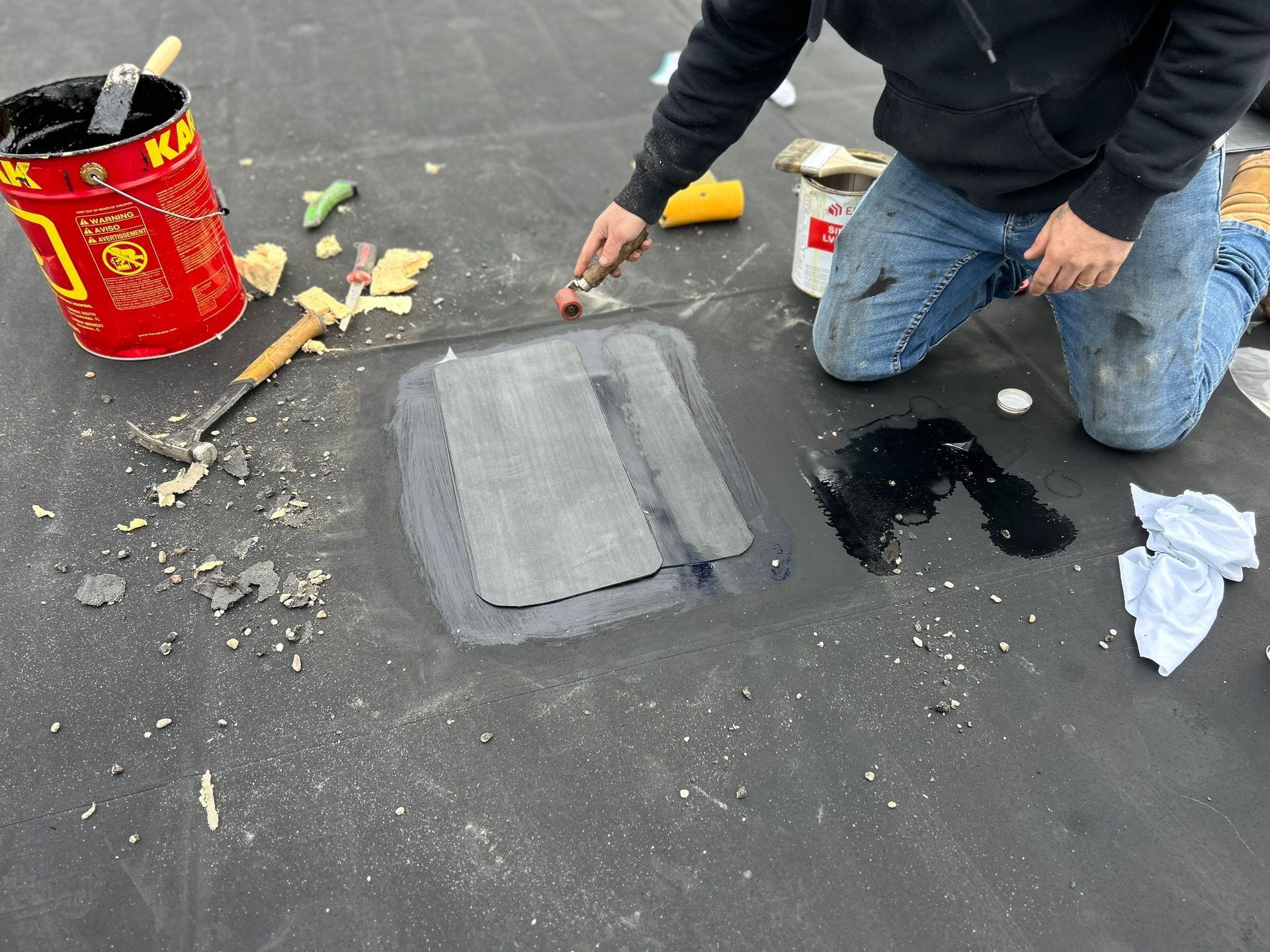 A person kneeling on a black roof surface, applying black adhesive to a patch over a repair area with a small roller.
