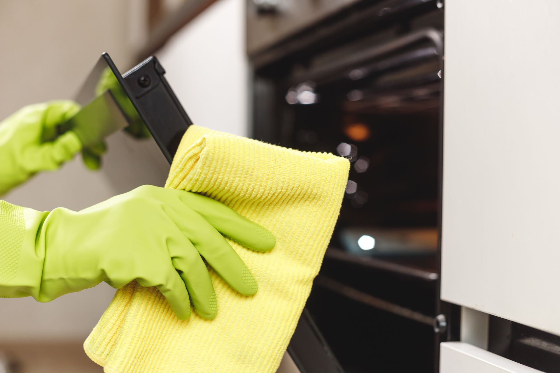 Green-gloved hands wiping a black oven with a yellow cloth in a kitchen-like setting