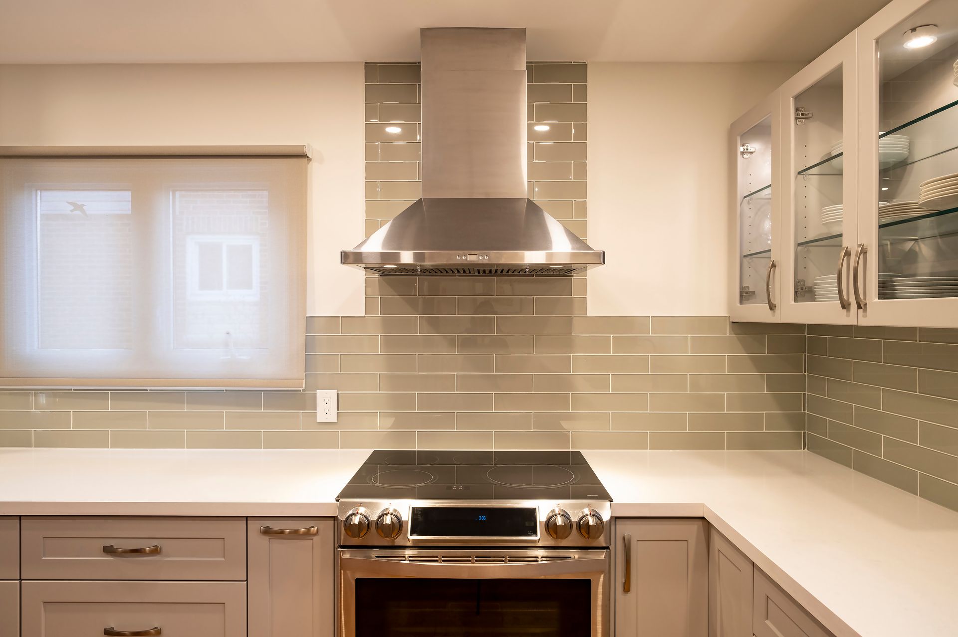 Modern kitchen with stainless steel range hood, beige tile backsplash, white countertops, and glass cabinets
