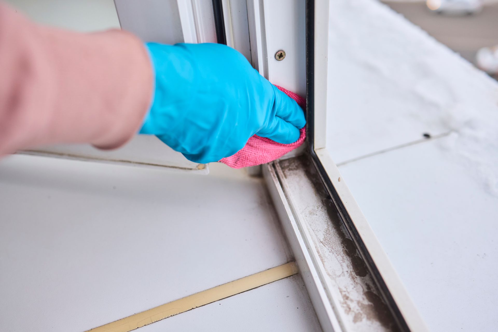 Gloved hand cleaning a window track with a pink brush and blue cloth