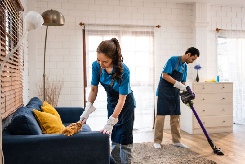 Two cleaners in blue uniforms vacuum and tidy a living room with a blue sofa and beige rug.