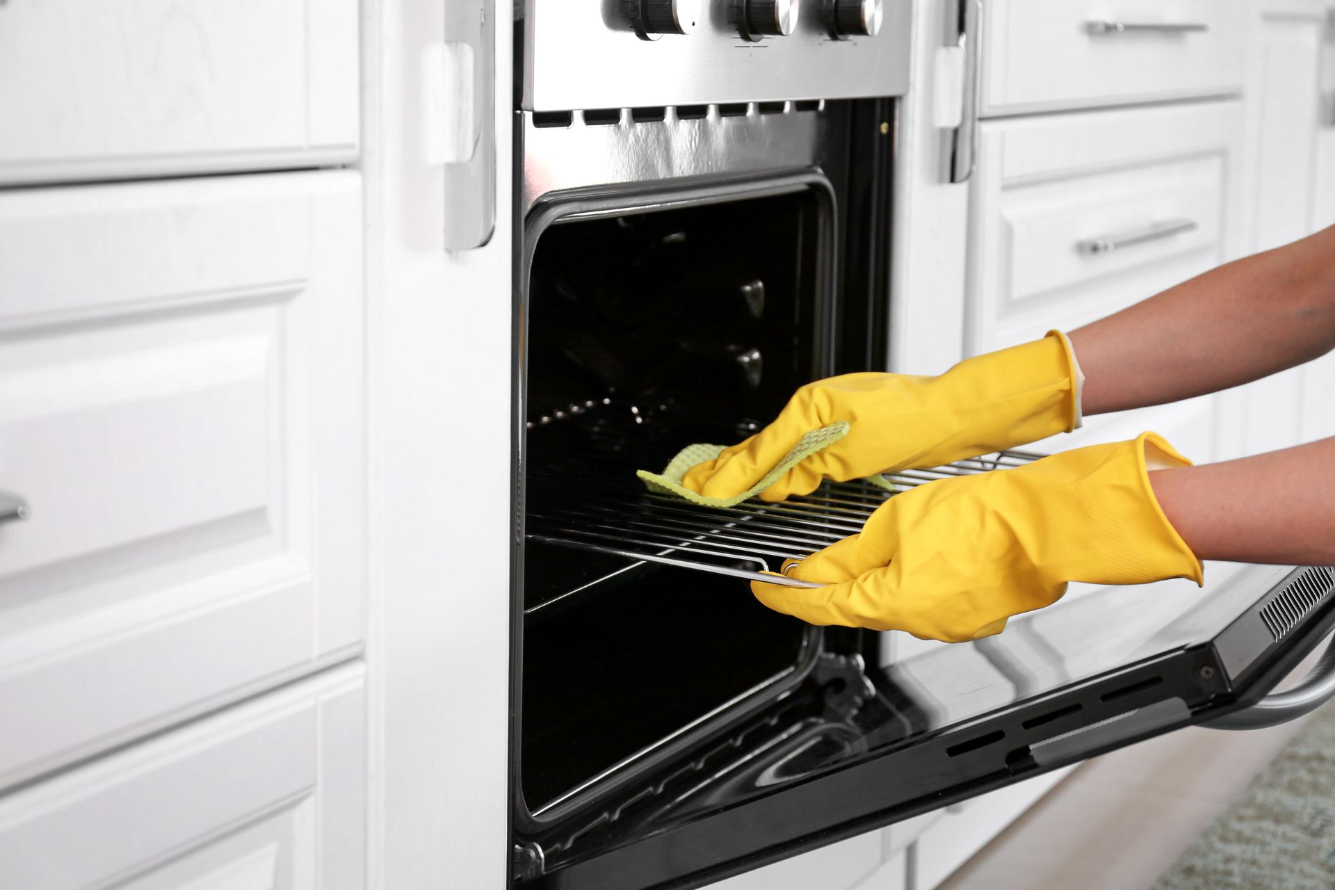 Yellow-gloved hands pulling a wire rack from a built-in oven in a white kitchen