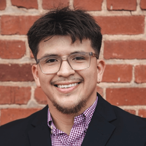 A man wearing glasses and a suit is smiling in front of a brick wall.
