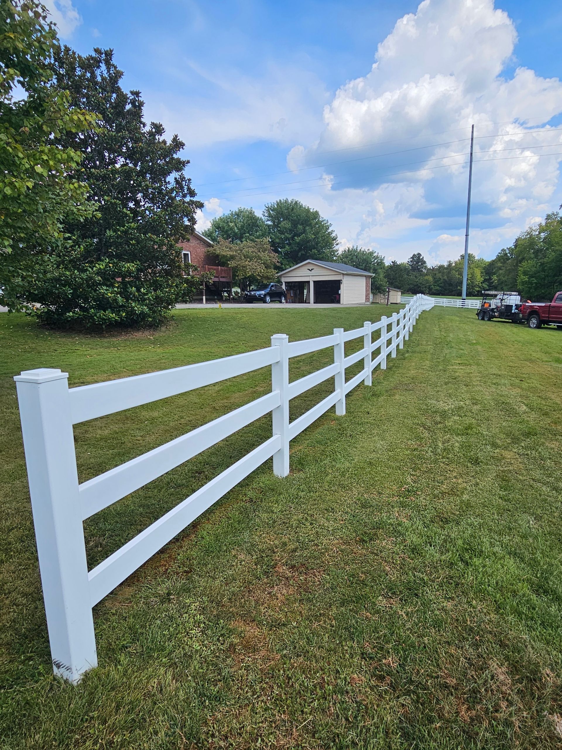 White fence along a grassy yard with trees, a small garage, and cars under a partly cloudy sky.