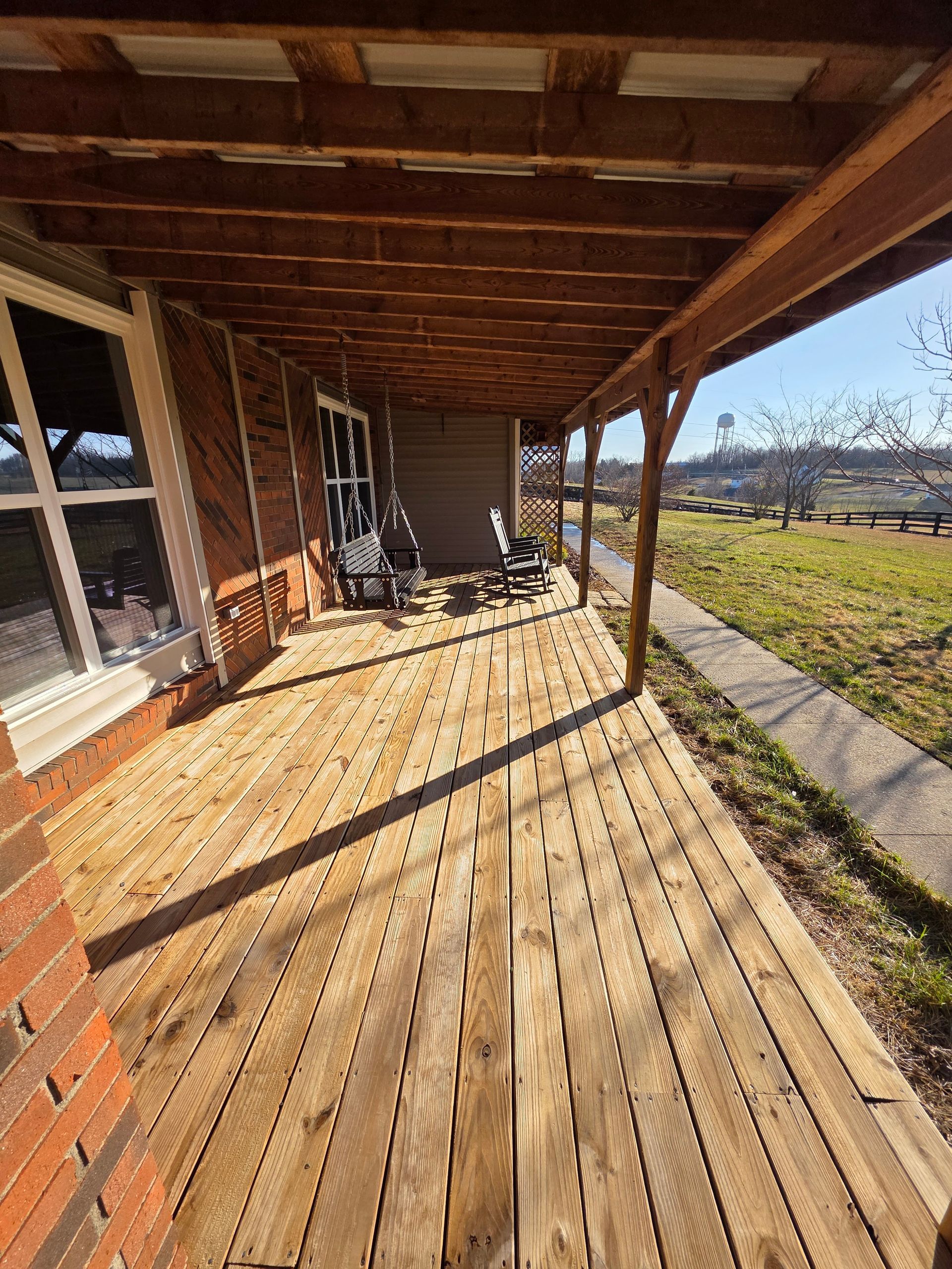 Sunlit wooden porch with railing beside a house, overlooking a grassy yard and open field.