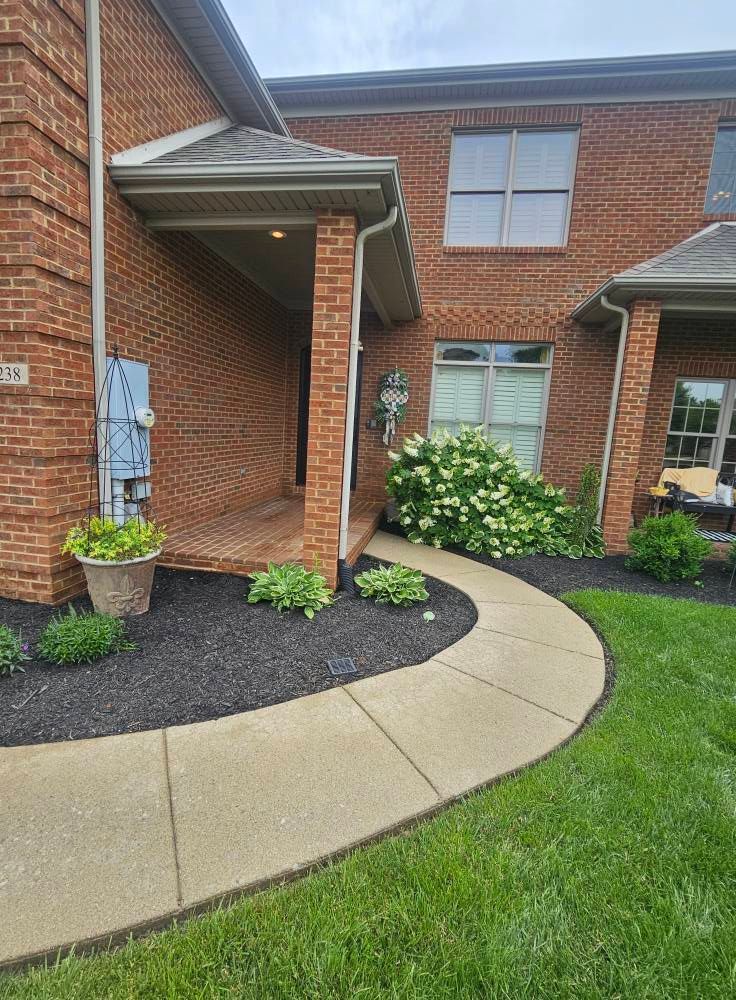 Curved concrete walkway beside brick townhouse patio with potted plants and flower beds