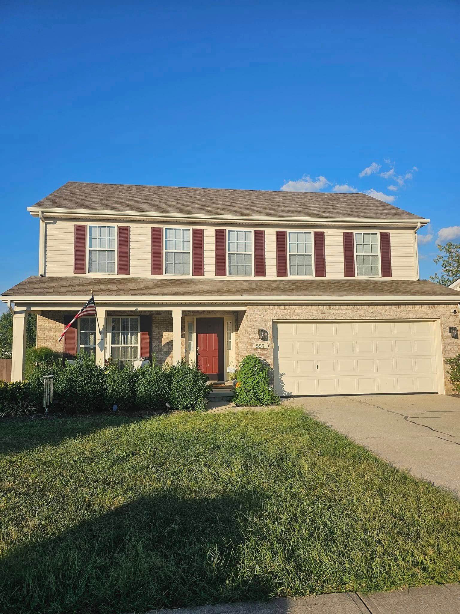 Two-story suburban house with beige siding, red shutters, a front lawn, and a two-car garage under a blue sky