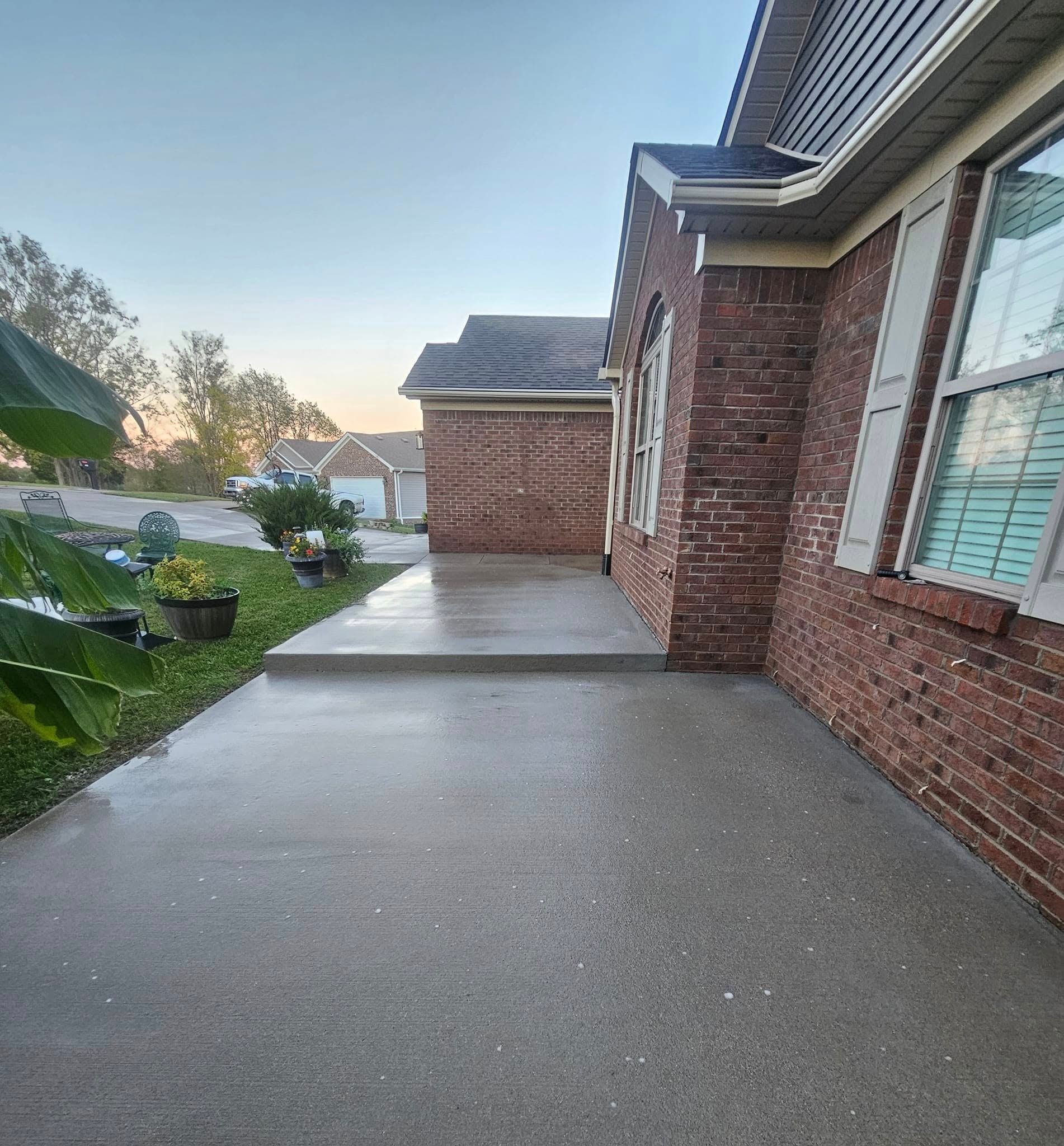 Brick house side walkway and driveway at dusk, with windows, lawn, and nearby homes.
