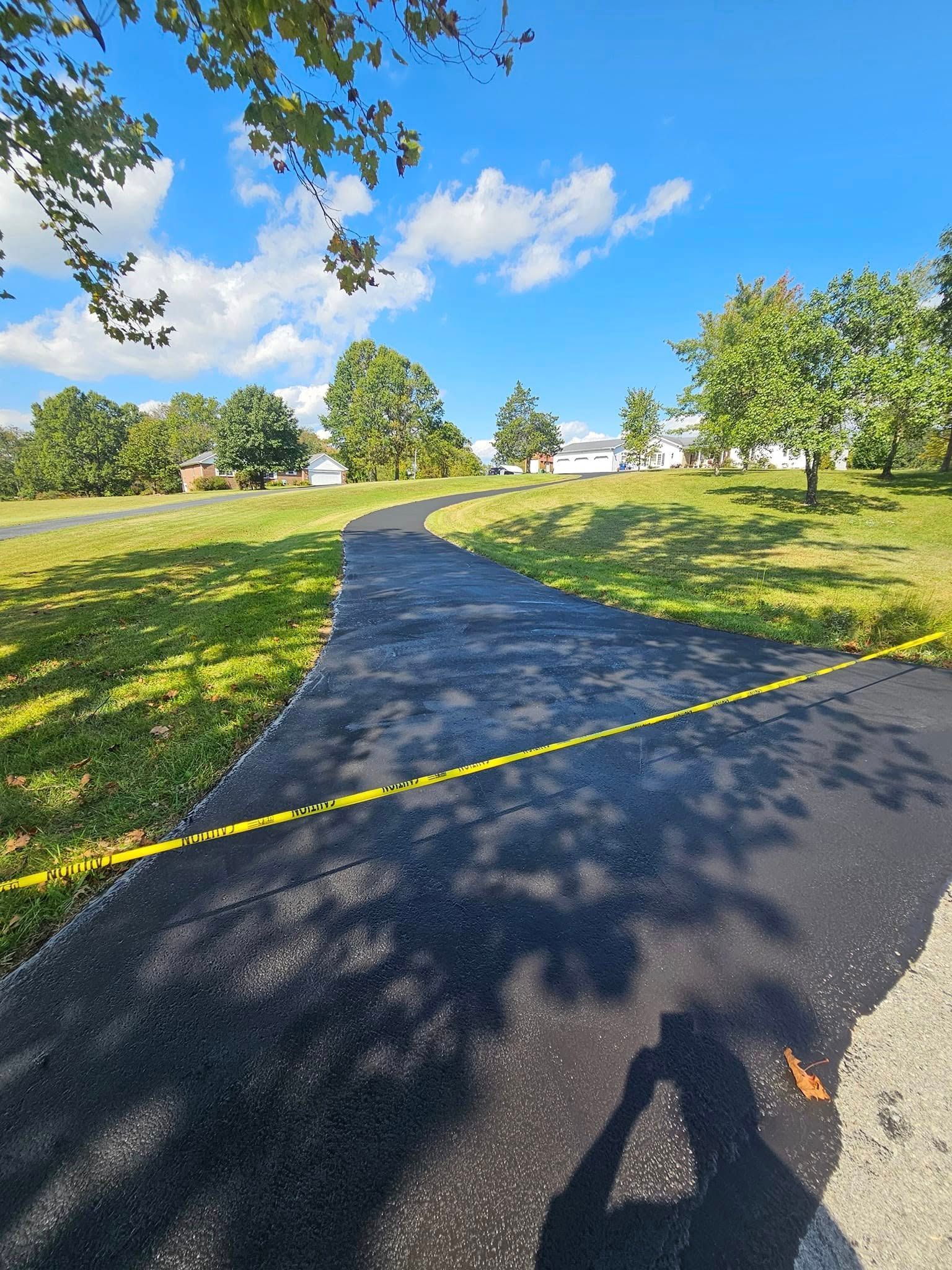 Sunlit park with a paved path, yellow tape across it, green grass, and trees under a blue sky.