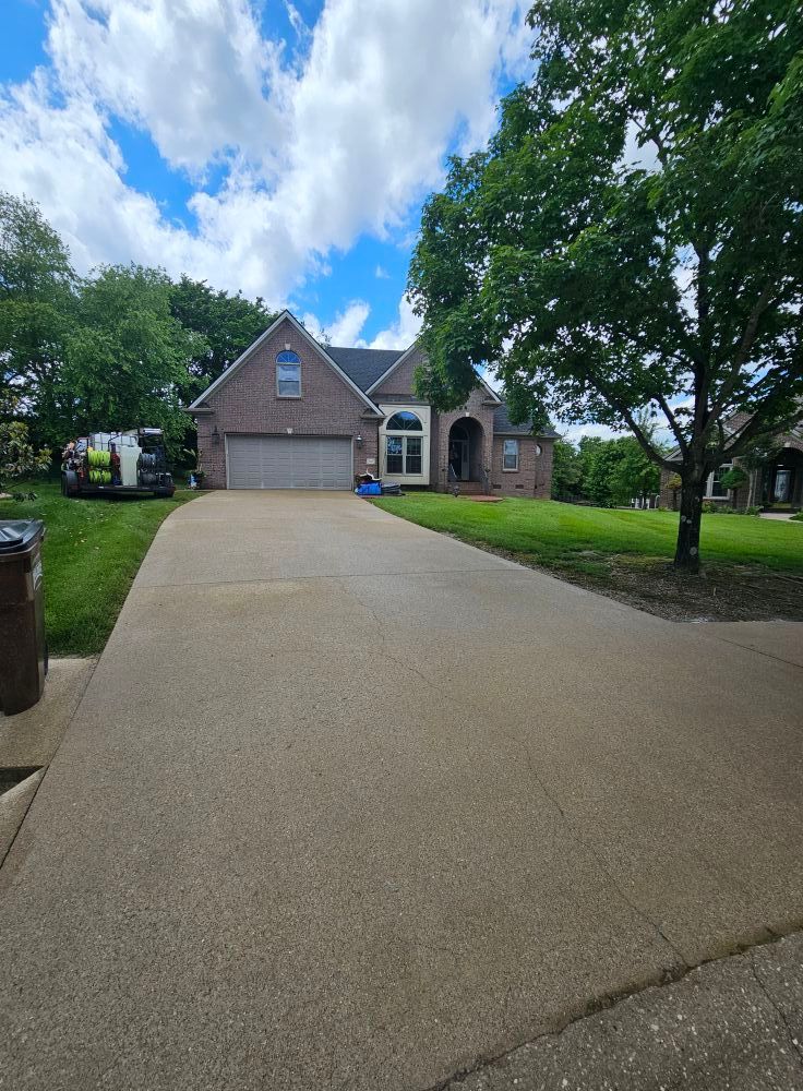Suburban house with a large concrete driveway, green lawn, and trees under a partly cloudy sky