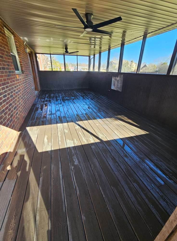 Covered wooden deck with a ceiling fan, brick wall, and sunlight casting shadows across the floor.