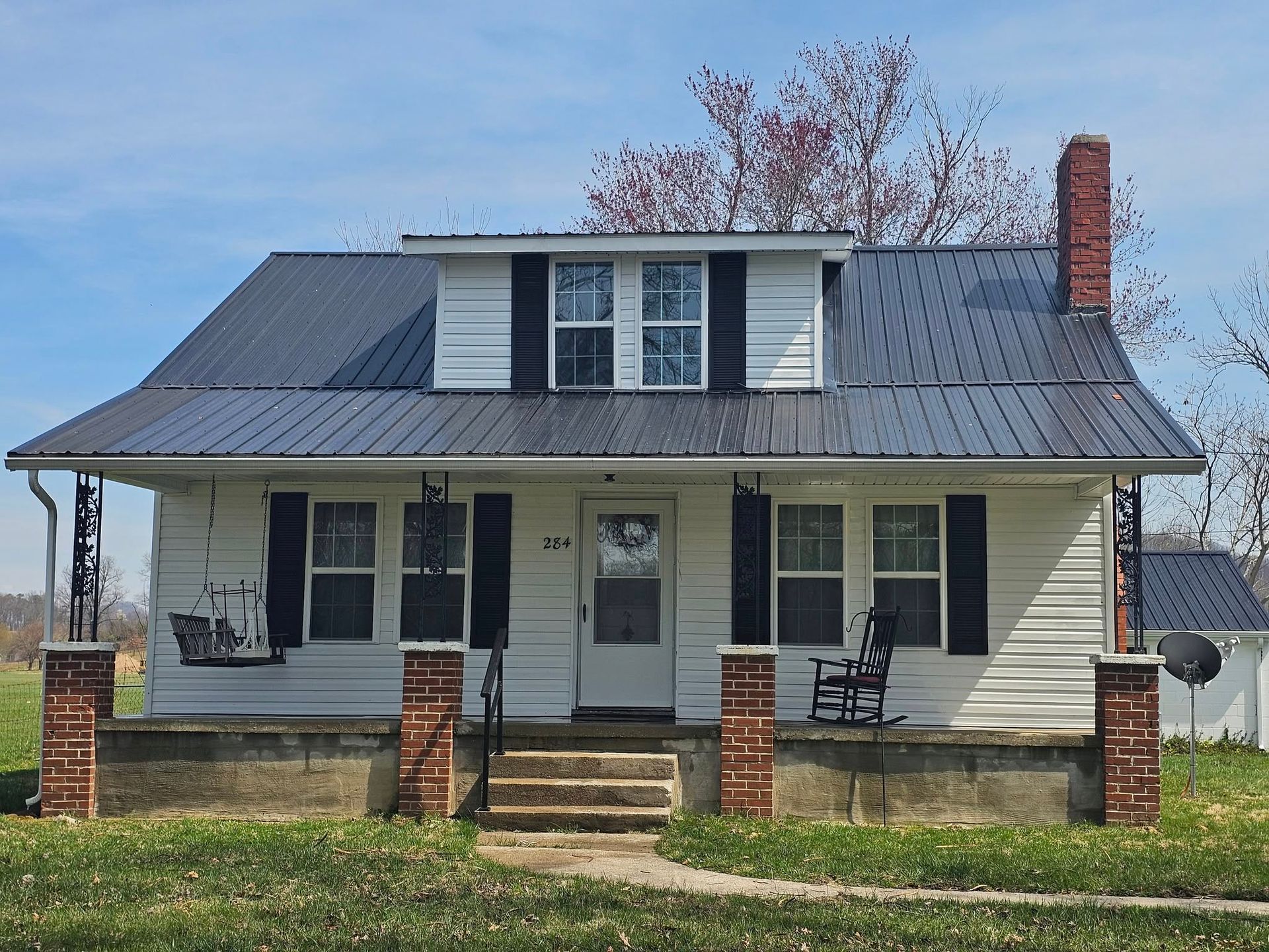 Small beige house with a metal roof, front porch, brick steps, and two chairs under a clear sky