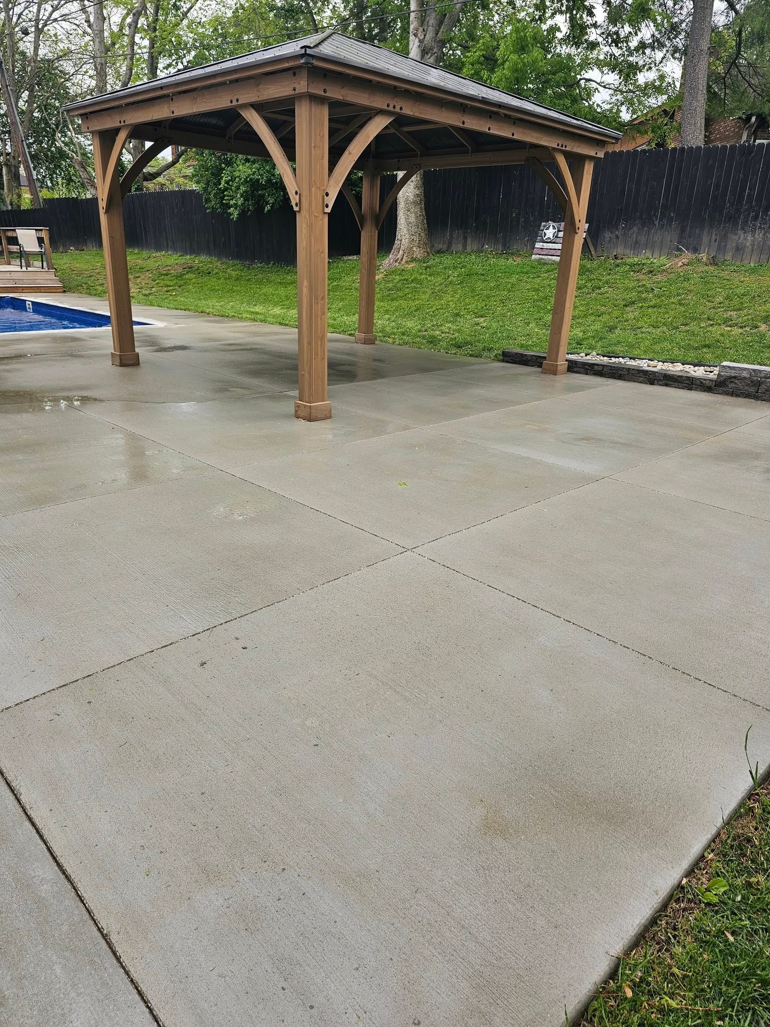 Wooden gazebo on a wet concrete patio beside grass and trees