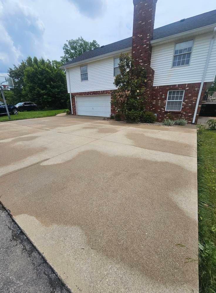 Residential driveway and two-story house with white siding, brick chimney, and patchy paved surface.