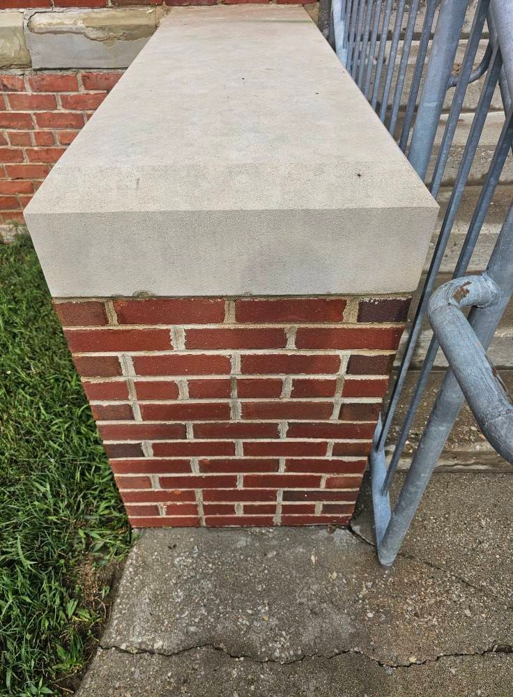 Brick pillar with a light concrete cap beside a metal gate and sidewalk.